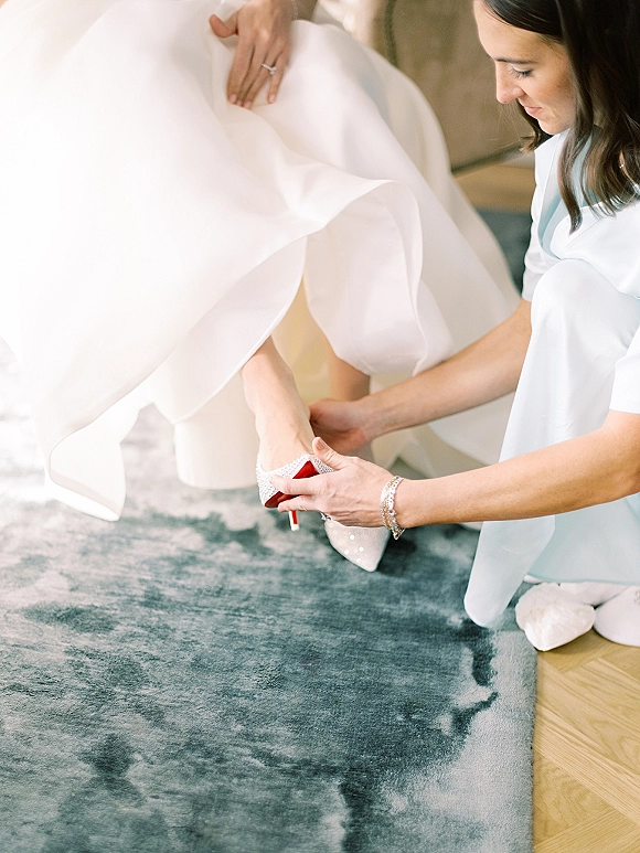Bridal shoes as the bride putting on shoes, sparkly white glitter heels with red soles on a blue rug, engagement ring and dress hem visible
