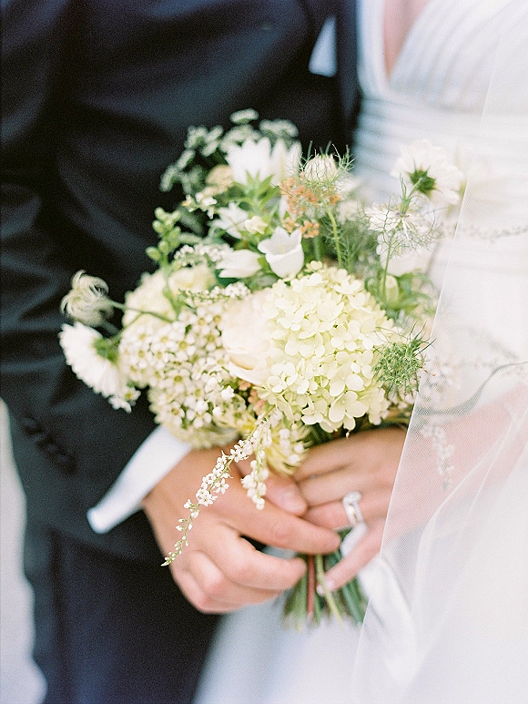 Bridal bouquet of white hydrangea and greenery held by the bride and groom, with her veil, his tuxedo, and rings in soft blur