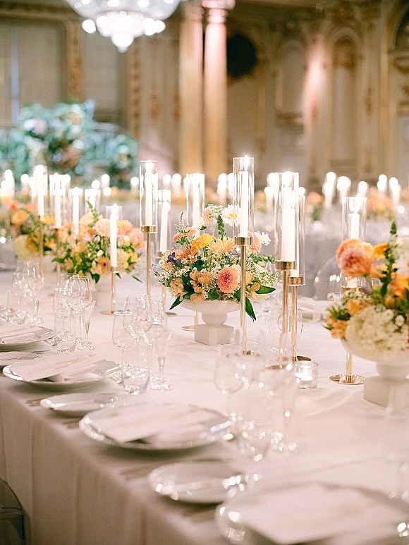 Reception tablescape with white wedding table setting, low florals and taper candles in gold holders, set under chandeliers in an ornate ballroom
