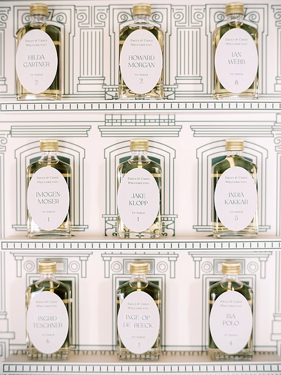 Wedding escort cards with mini liquor bottles, custom oval name labels, and gold caps arranged on a shelf against a white backdrop with green line art
