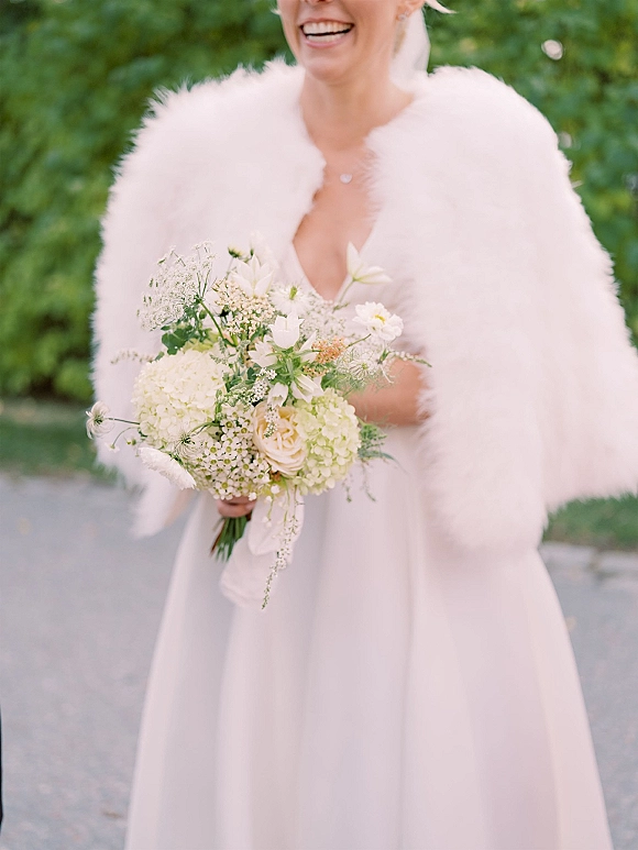 Bride portrait holding bouquet, laughing in a strapless wedding dress with a white fur shawl on an outdoor path lined with green trees