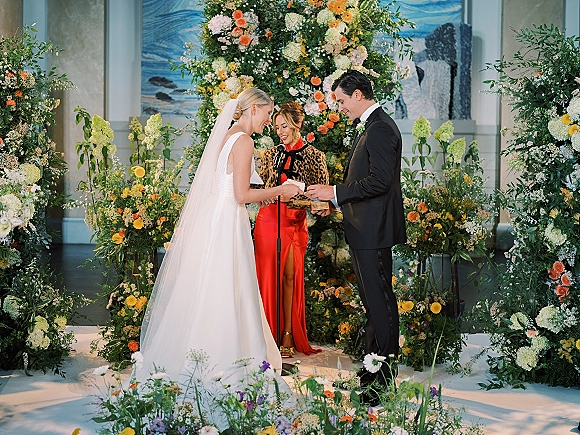 Wedding vows as bride and groom stand with officiant at floral arch, bride’s long veil and microphone in an indoor venue