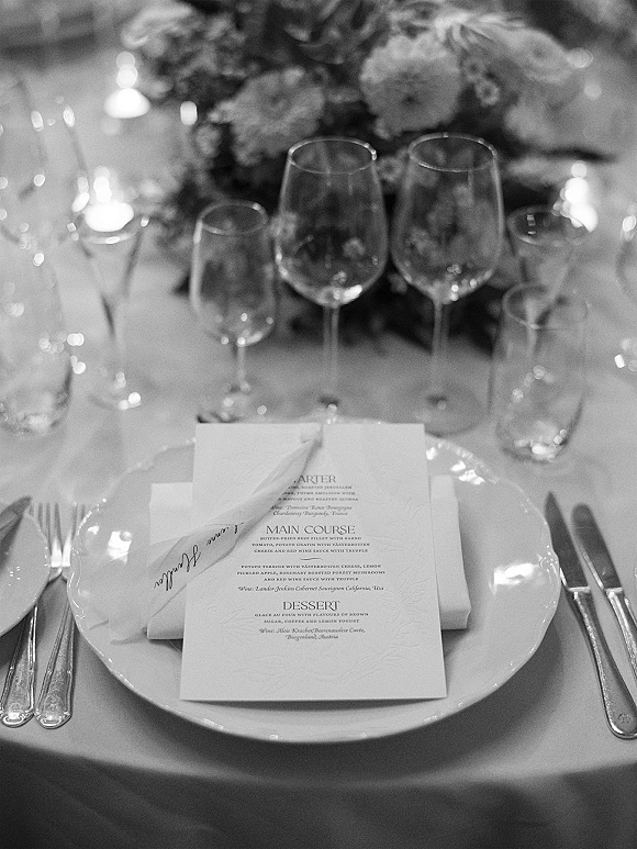 Wedding tablescape with wedding place setting featuring menu card on plate, folded napkin, candles, floral centerpiece, and soft bokeh lights