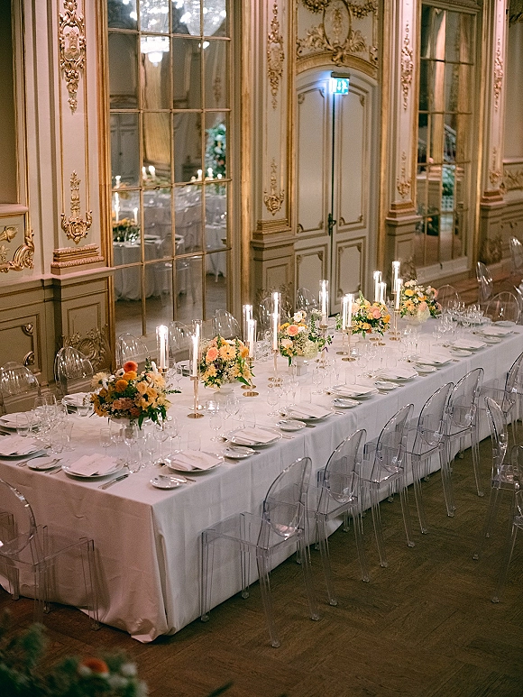 Reception tablescape on a long banquet table with white tablecloth, tall glass candles, orange and yellow florals, and clear acrylic chairs in a gilded ballroom with mirrors