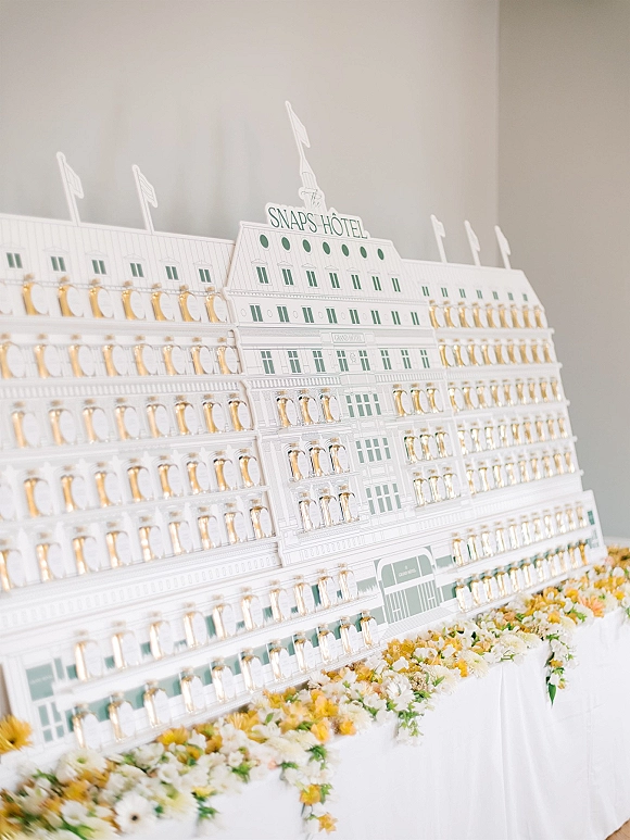 Champagne wall with wedding champagne wall flutes, a white tablecloth and yellow-white floral garland against a minimal white backdrop
