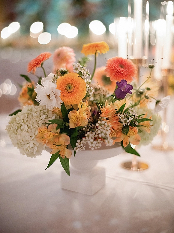 Wedding centerpiece with a mixed floral arrangement of orange blooms, white hydrangea and greenery in a compote vase, taper candles behind on a bokeh-lit reception table