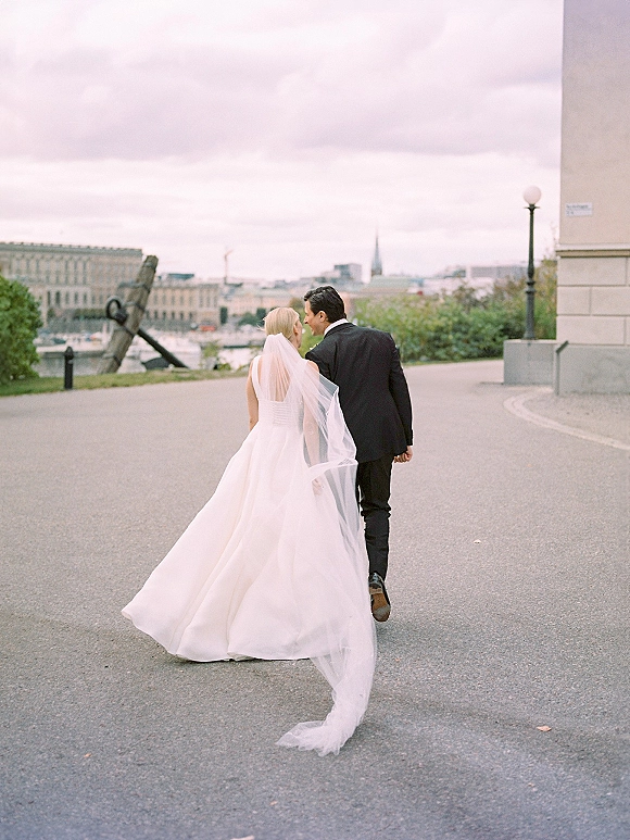 Couple portrait of bride and groom walking away holding hands, her long cathedral veil and dress train flowing on an urban street skyline