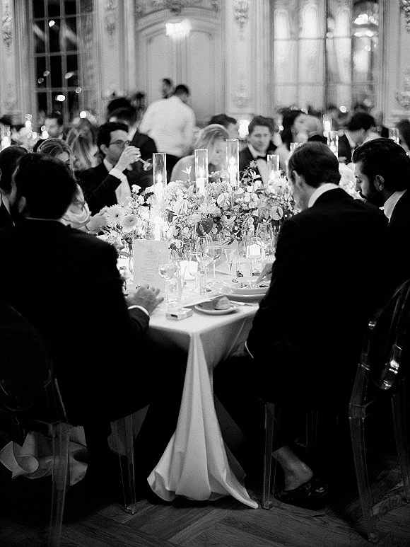 Wedding reception dinner with round banquet tables, candle and floral centerpieces, and guests in tuxedos inside an ornate ballroom with tall windows