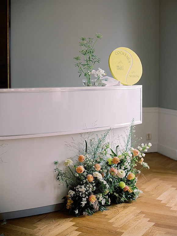 Wedding bar decor with wedding cocktail bar setup, bud vases and floral arrangement beside a cocktail sign on a bar counter against a gray wall