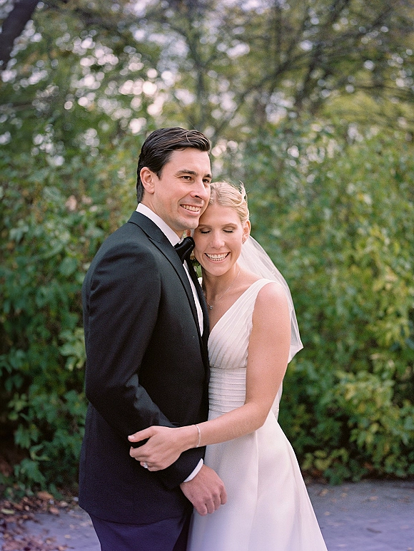 Couple portrait of bride and groom hugging, her veil draped over a simple wedding dress as he wears a tuxedo on a green path