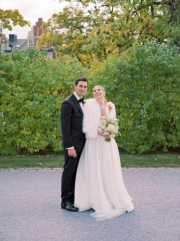 Couple portrait of bride and groom standing on a garden path, bride holding a white bouquet in gown and faux fur stole, groom in tuxedo