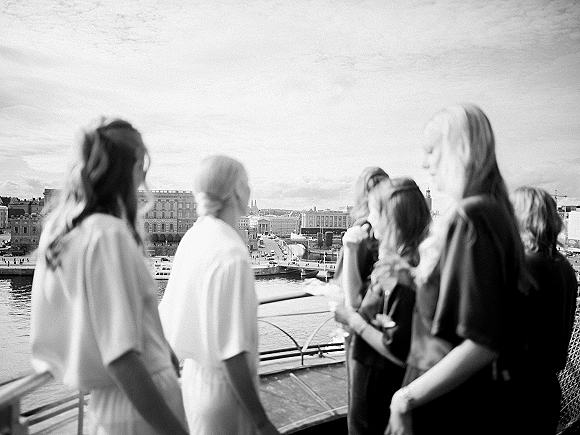 Wedding guests in dresses and short sleeve shirts lean on a railing, watching the river with city skyline and bridge under cloudy sky