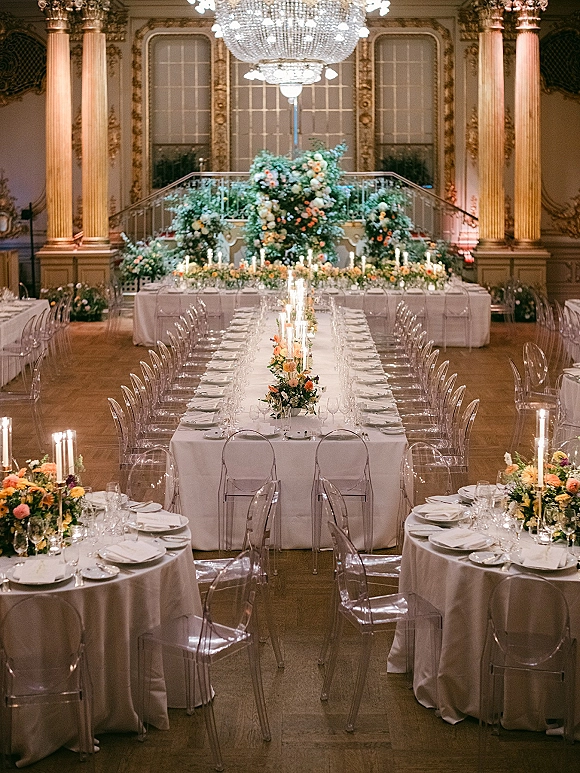 Reception tablescape with long head table decor, taper candles and floral centerpieces, clear acrylic chairs in an ornate grand ballroom with chandeliers