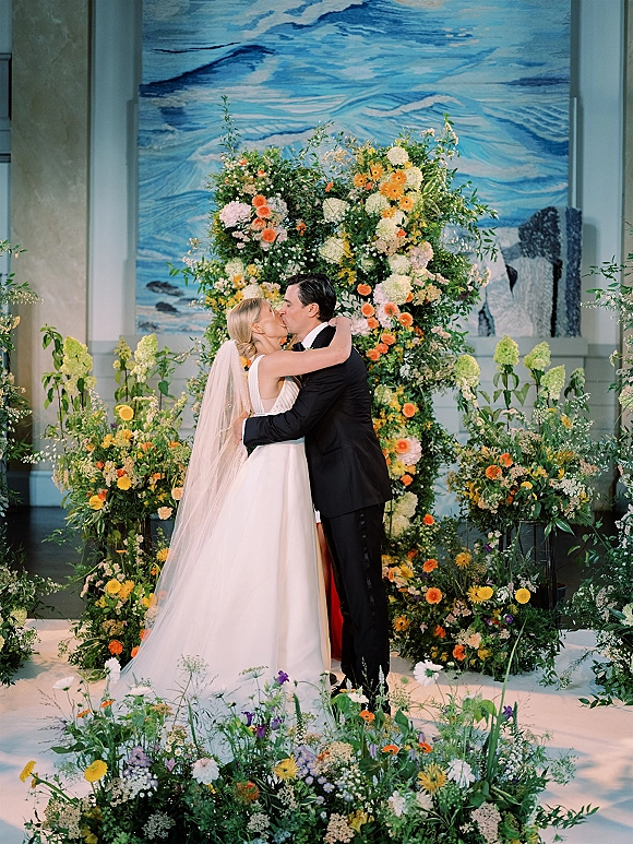 Wedding kiss as bride and groom embrace beneath a wildflower floral arch, veil flowing, with a painted mural backdrop at the indoor altar