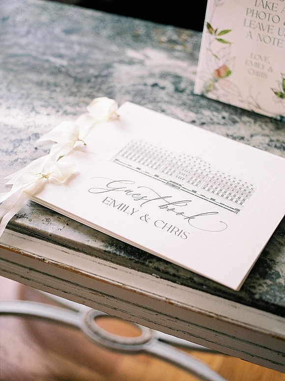 Wedding guest book with guest book sign script lettering, satin ribbon bows, and greenery illustration on a marble tabletop near drawer handle