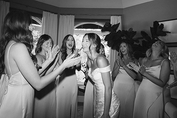 Bridal party moment as bride laughs while bridesmaids cheering bride and clapping in a bright bridal suite with arched windows and curtains