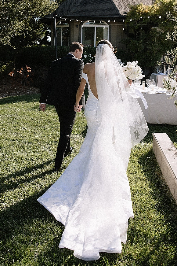 Newlyweds walking away hand in hand, bride in strapless gown with long veil and bouquet beside groom in black tux under string lights on patio lawn