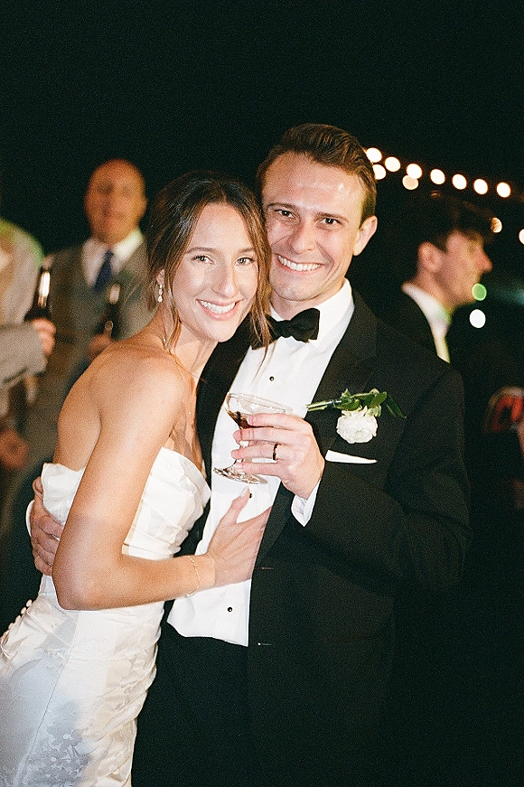 Couple portrait of bride and groom hugging, smiling in black tie attire with cocktails under string lights at a nighttime reception