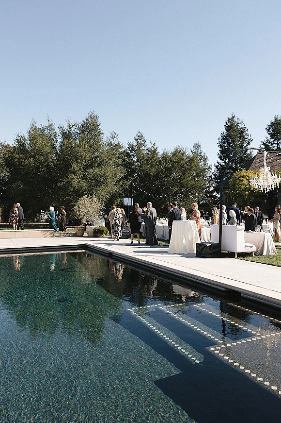 Wedding cocktail hour poolside wedding reception setup with white high-top tables, lounge seating, chandelier and string lights by the pool deck