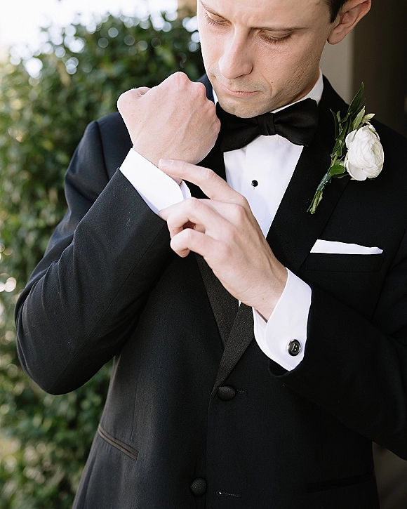 Groom portrait with groom adjusting cufflinks on a black tuxedo and bow tie, white rose boutonniere against outdoor greenery backdrop