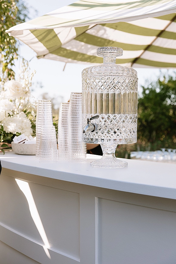 Drink dispenser crystal drink dispenser with spigot on a white bar counter, stacked plastic cups and floral decor under striped umbrella outdoors