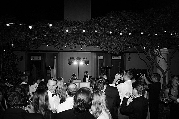 Wedding reception dance with guests in tuxedos cheering on a crowded outdoor courtyard dance floor under string lights by the DJ booth at night