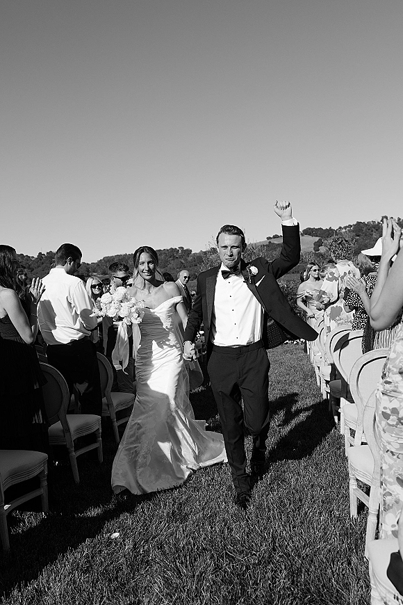 Wedding recessional as newlyweds walk hand in hand up an outdoor aisle, bride with bouquet, groom in tux fist pump, guests cheering on lawn