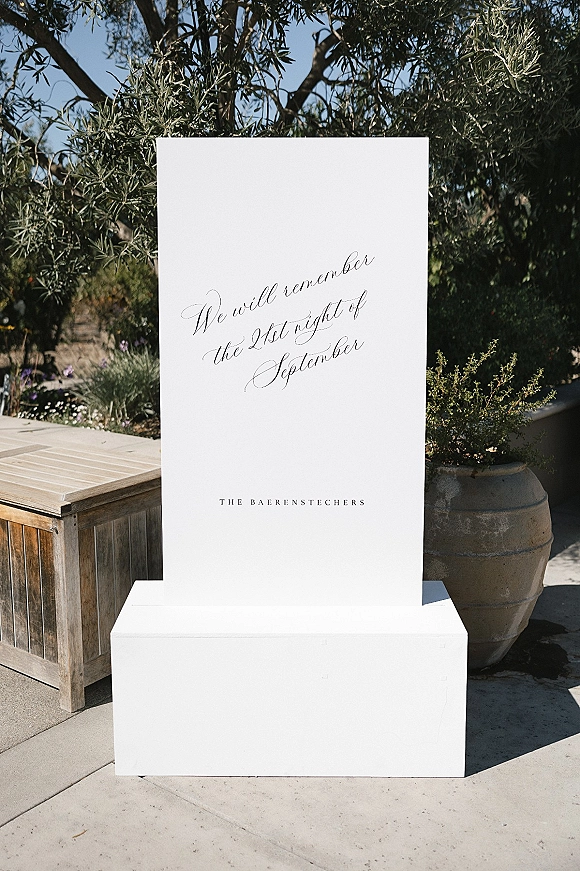 Wedding welcome sign with calligraphy wedding sign lettering on a white board stand, set in an outdoor courtyard by an olive tree and bench