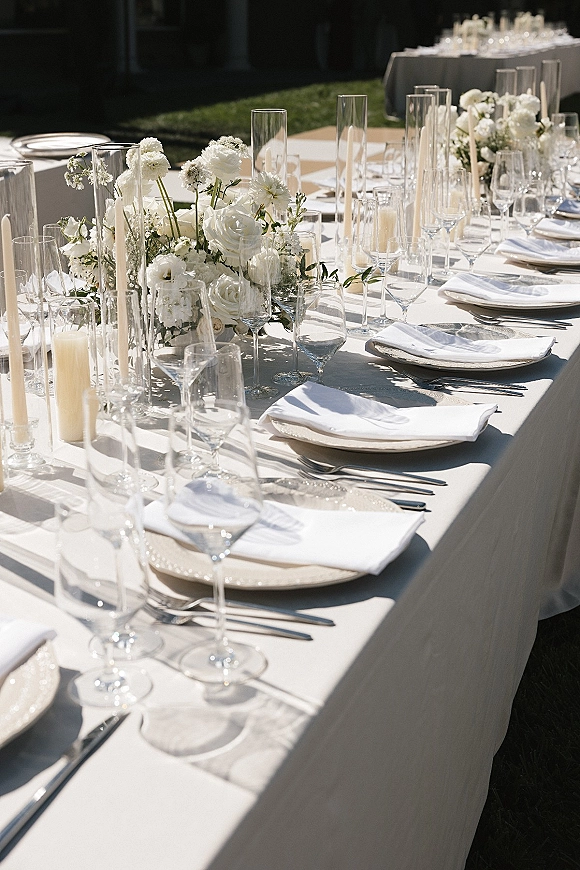 Reception tablescape with white floral centerpiece, taper candles and glass cylinder vases, set with crisp napkins and glassware on an outdoor lawn