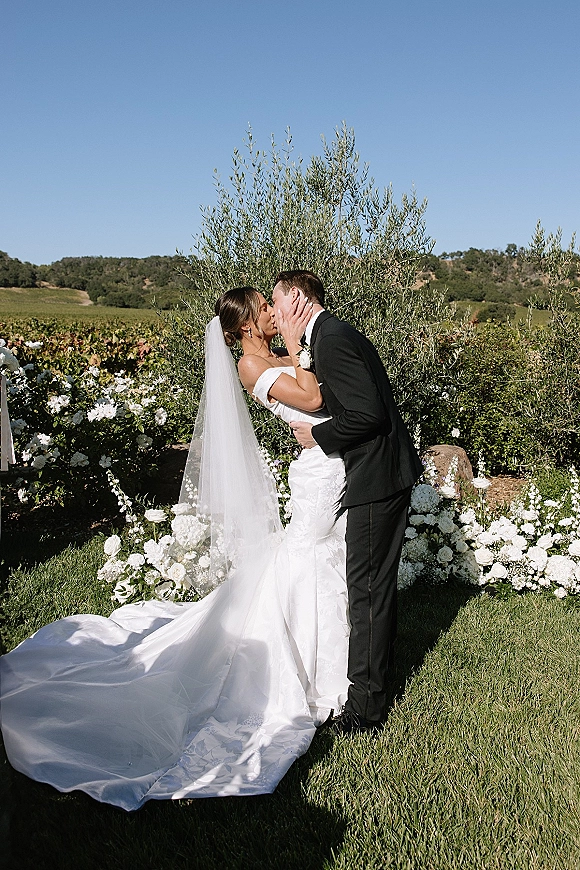 Wedding kiss as bride and groom kiss in side profile, her cathedral veil and bouquet trailing in a sunny vineyard garden with hills
