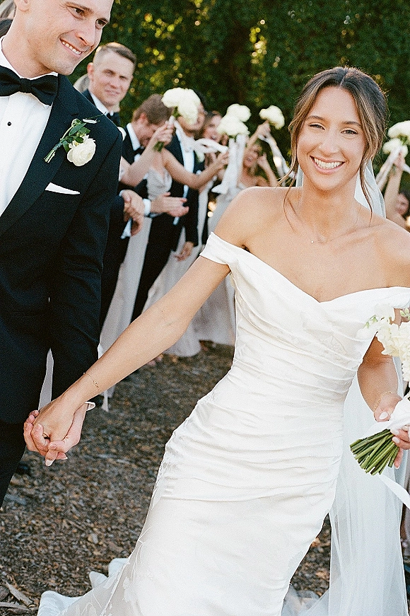 Wedding recessional as bride and groom walking hand in hand, bride smiling with white rose bouquet, wedding party lining a garden path