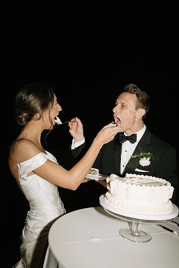 Wedding cake cutting as the bride feeding groom cake from a white frosted slice on a glass stand, posed against a dark backdrop