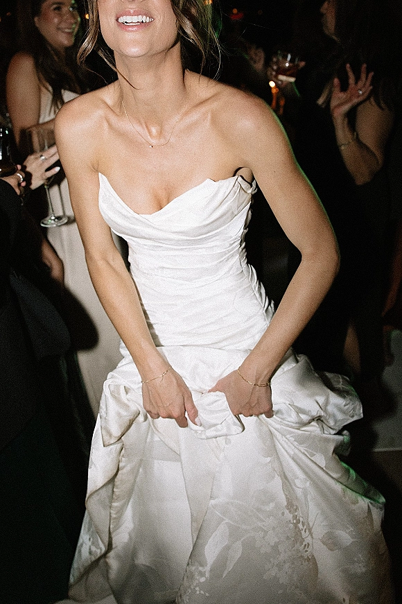 Bride dancing in a strapless ruched wedding dress at a dim wedding reception, holding her skirt as guests with wine glasses cheer behind her