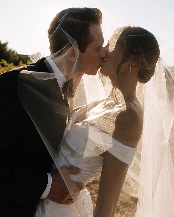 Wedding kiss portrait of bride and groom kissing under a flowing veil at sunset, groom in tuxedo with bow tie and boutonniere