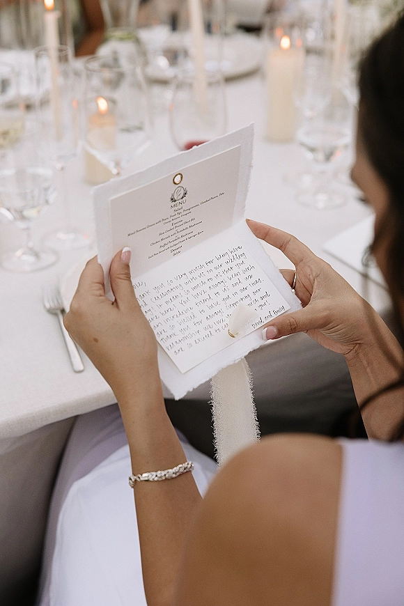 Wedding menu card with a handwritten note tied with ribbon at a candlelit reception place setting with glassware and blurred guests behind