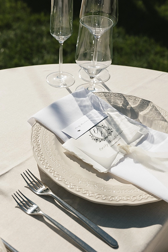 Wedding tablescape with a white linen napkin and monogram menu card tied with ribbon on a charger plate, with wine glasses and greenery backdrop
