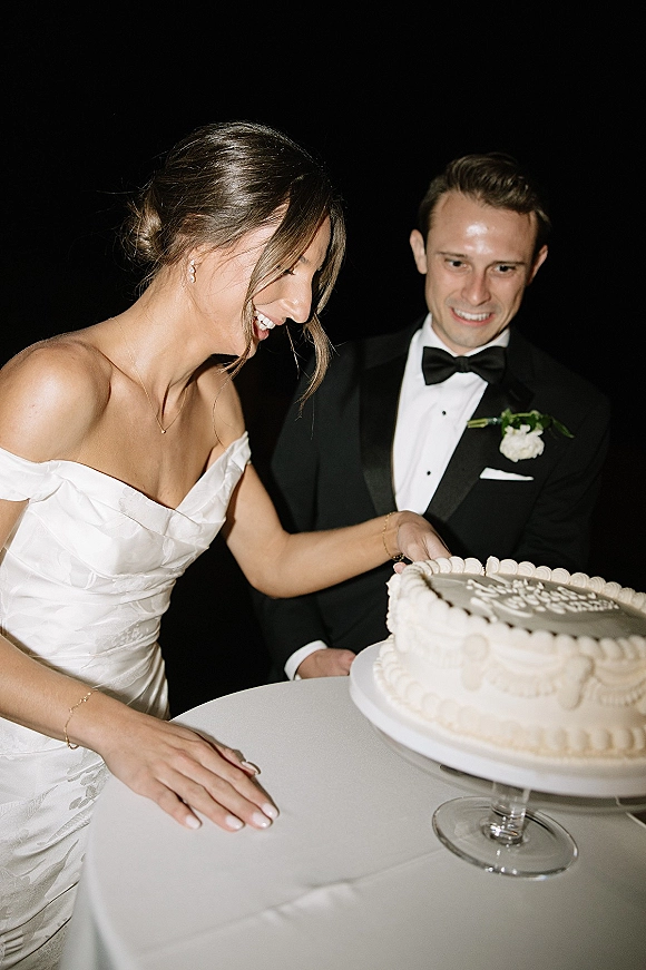 Wedding cake cutting as bride in strapless dress and groom in tuxedo slice a single-tier white buttercream cake on glass stand, dark backdrop