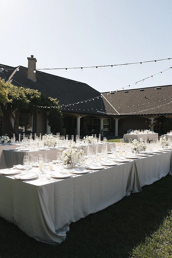 Reception tablescape for an outdoor wedding reception with long white linen banquet tables, white florals in cylinder vases, candles, and string lights overhead