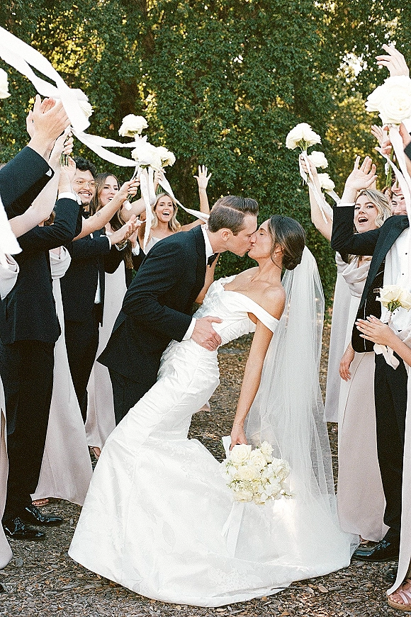 Wedding kiss portrait of the couple in a dip just married kiss as friends cheer with ribbon streamers, white rose bouquets, on a garden path