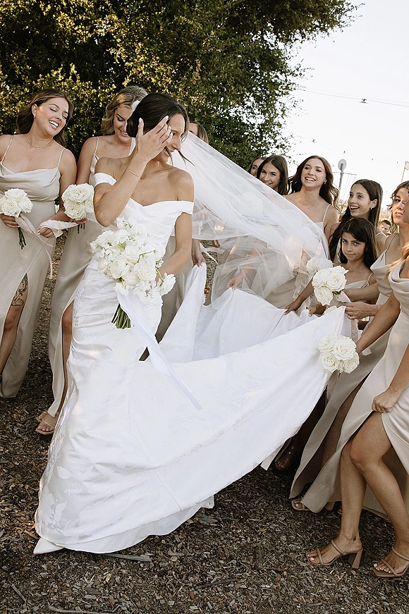 Bride with bridesmaids holding her long wedding veil as she carries a white rose bouquet on a tree-lined path with string lights overhead