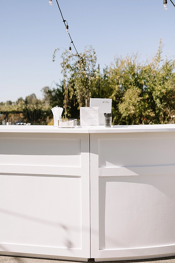 Wedding bar setup with white bar counter, drink stirrers and cocktail napkins in an acrylic tray under string lights with trees and blue sky