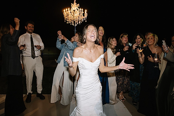 Wedding dance party with bride dancing reception in an off-the-shoulder dress as guests cheer holding champagne glasses under a chandelier indoors