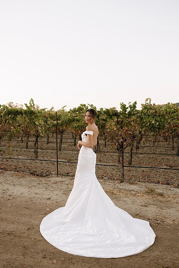 Bridal portrait in an off the shoulder wedding dress, satin gown with long train, bride looking back on a vineyard path under open sky