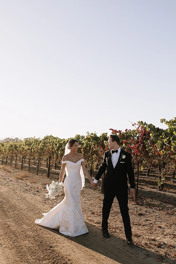 Couple portrait of bride and groom holding hands, walking a vineyard path at golden hour, her veil and white rose bouquet beside his tuxedo
