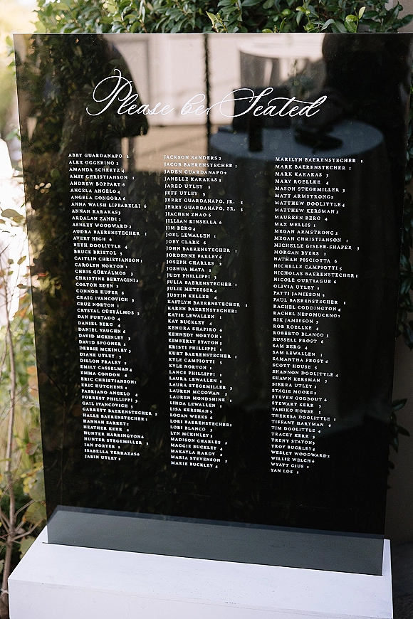 Wedding seating chart acrylic seating chart with calligraphy guest list on a white pedestal stand, set outdoors amid lush greenery