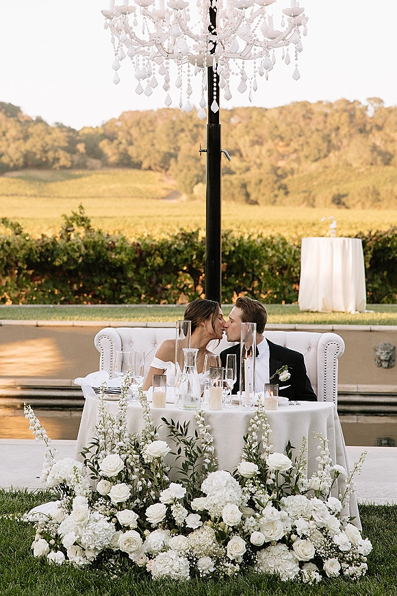 Wedding kiss portrait of bride and groom kissing at a candlelit sweetheart table with white roses, hydrangeas, and a crystal chandelier in a vineyard
