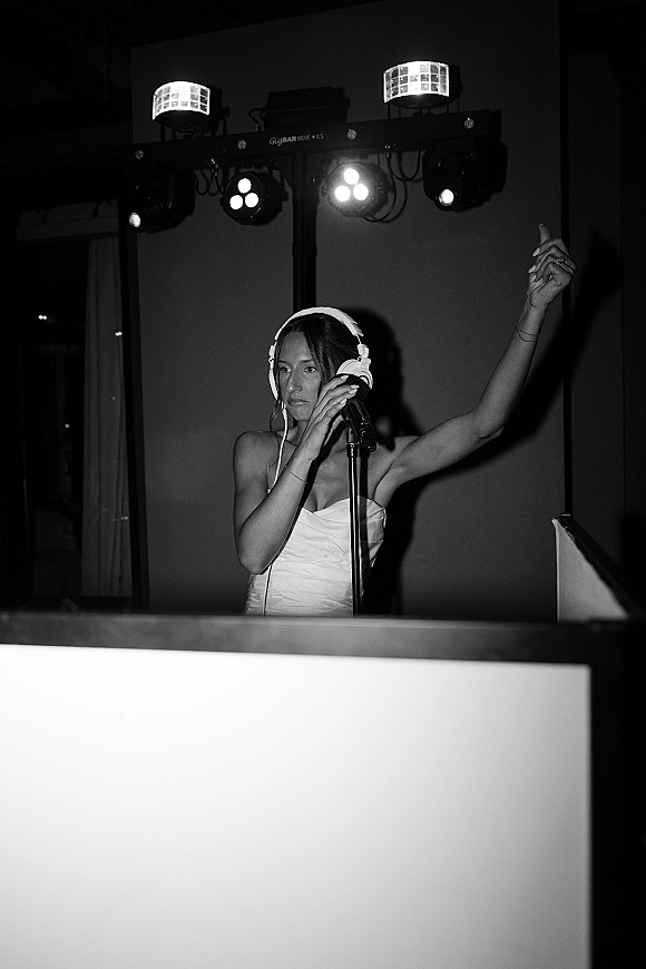 Wedding DJ moment with the bride DJing behind a booth, wearing headphones and holding a microphone under stage lighting indoors