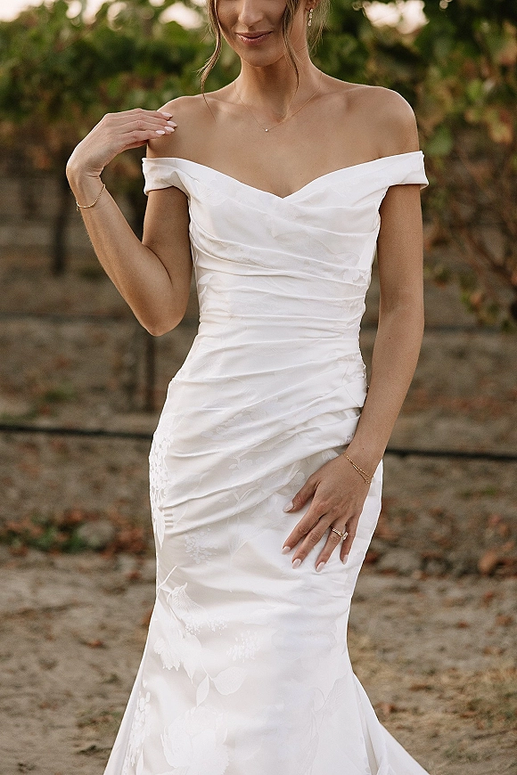 Wedding dress in ruched satin with an off the shoulder neckline, floral jacquard, and minimal jewelry, photographed in a vineyard path