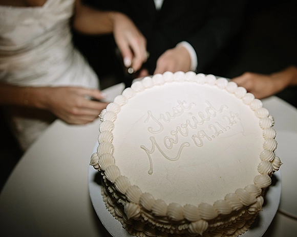 Wedding cake cutting as bride and groom use a cake knife on a white buttercream cake with piped border and inscription at a reception table