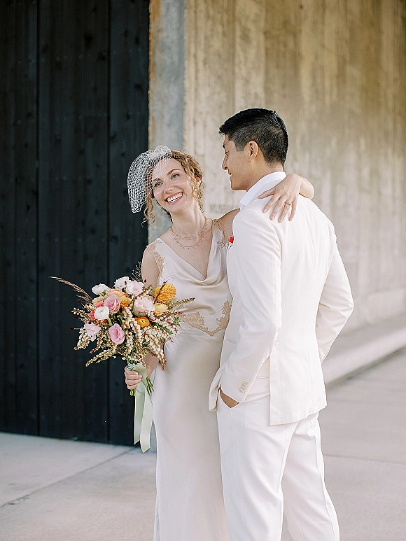 Couple portrait of bride and groom embrace, her birdcage veil and colorful bouquet with ribbon against a concrete wall backdrop